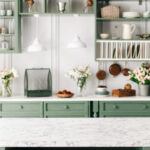 Clean and empty marble countertop, green vintage kitchen furniture with lots of flowers and bowl of strawberries, pair of white hanging pendant lights, various crockery in blurred background